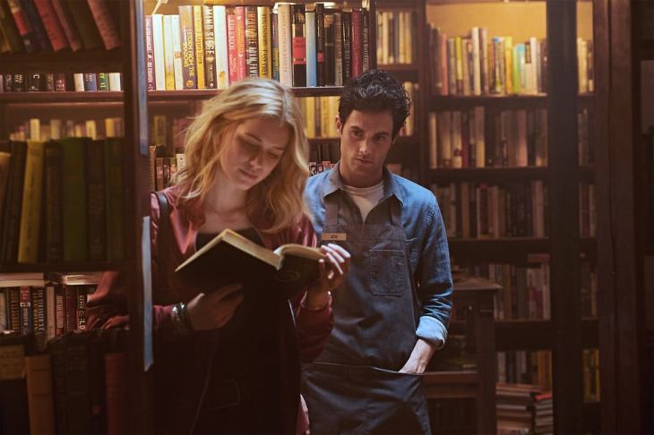 A still from series 'You' on Netflix. A young man peers over a young woman's shoulder as she reads in a bookshop.