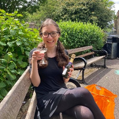 Me celebrating in the park after my exams. I hold a glass of prosecco and sit on a bench.