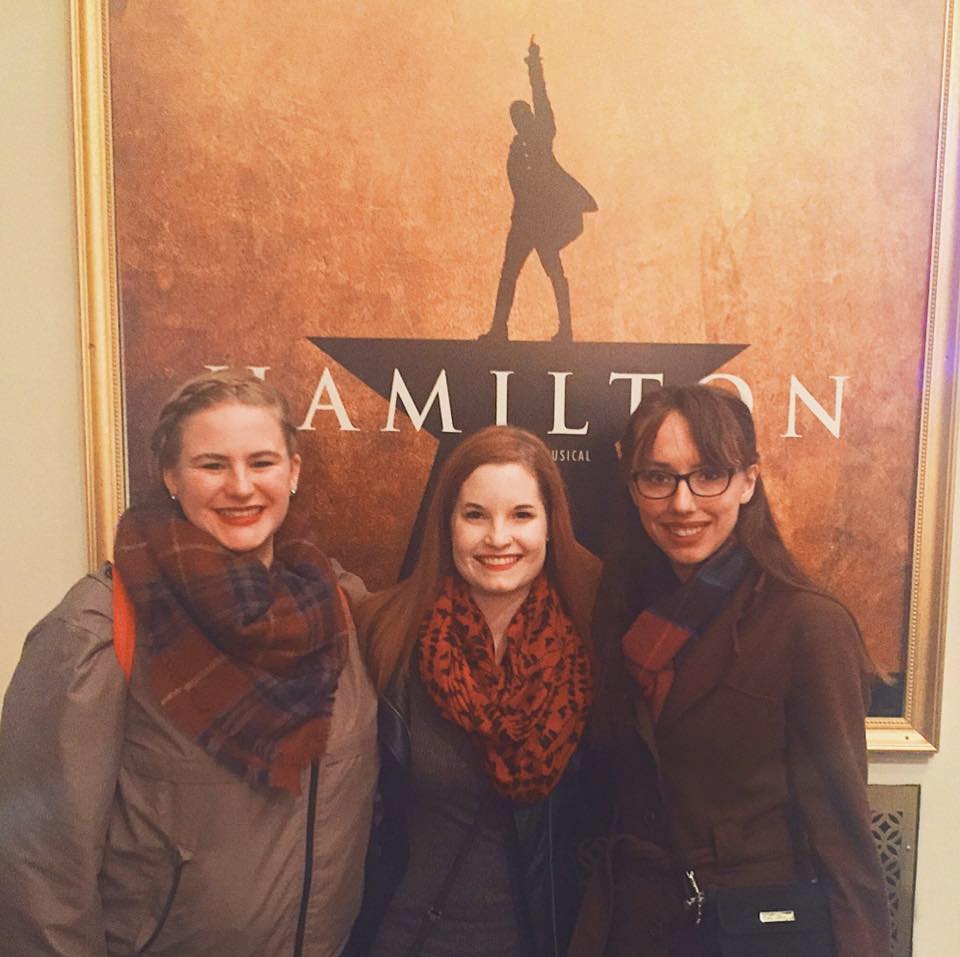 Three friends stand together in front of a poster for 'Hamilton'.