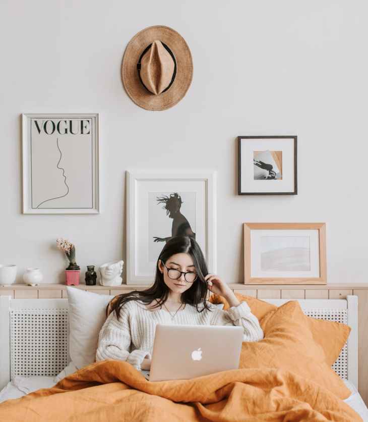 A woman sits in bed and uses her laptop under the covers. The background is covered in posters and artwork.