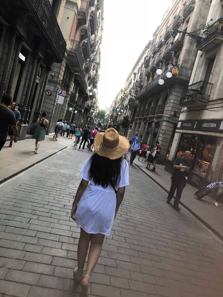 A young woman walks down a cute street with her back to the camera, wearing a straw hat.