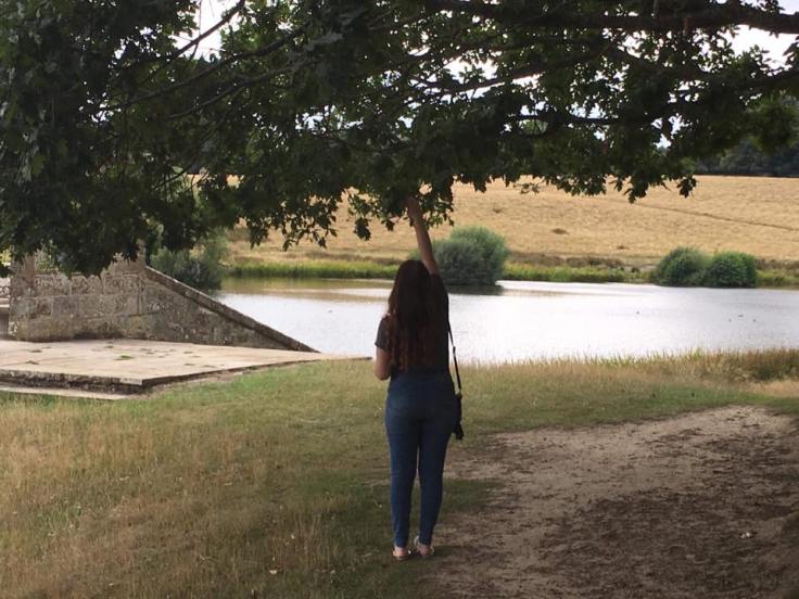 A young woman stands on a path covered with grass, under a tree.