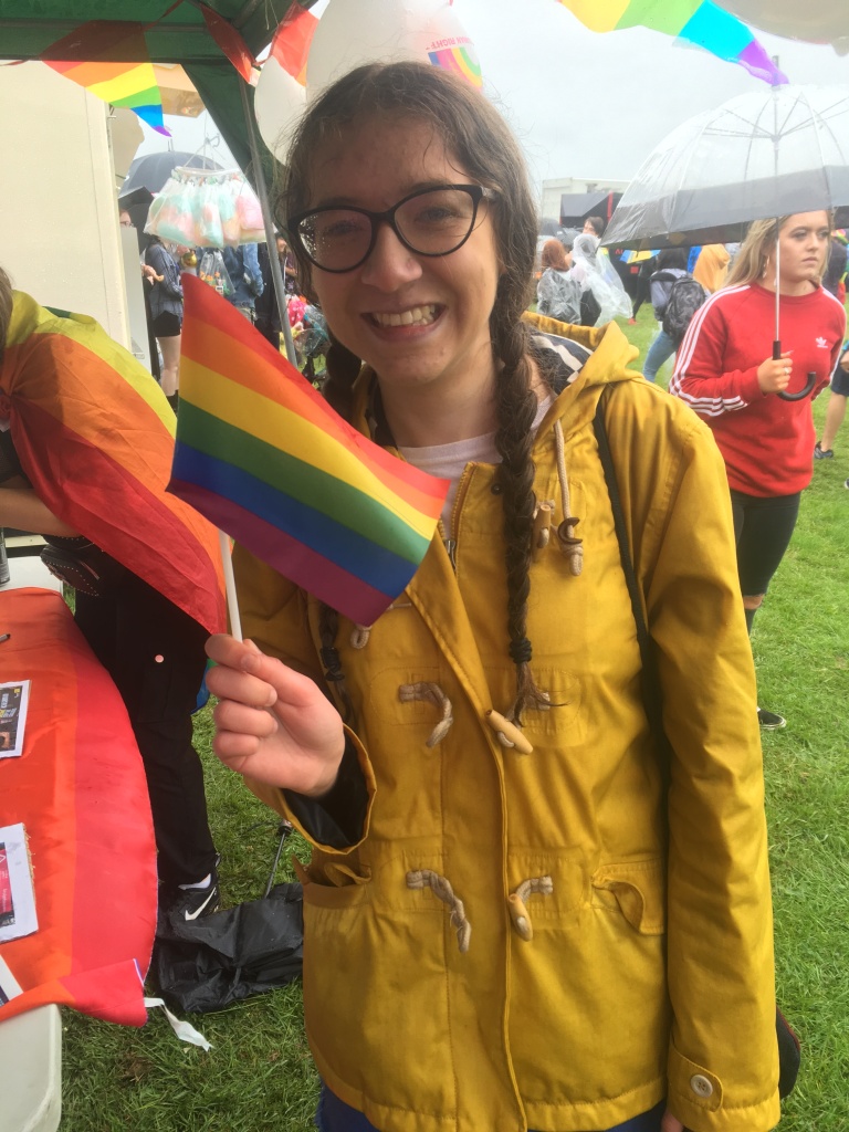 I'm standing with a small Pride flag in a yellow rain-coat. I have been rained on a lot!