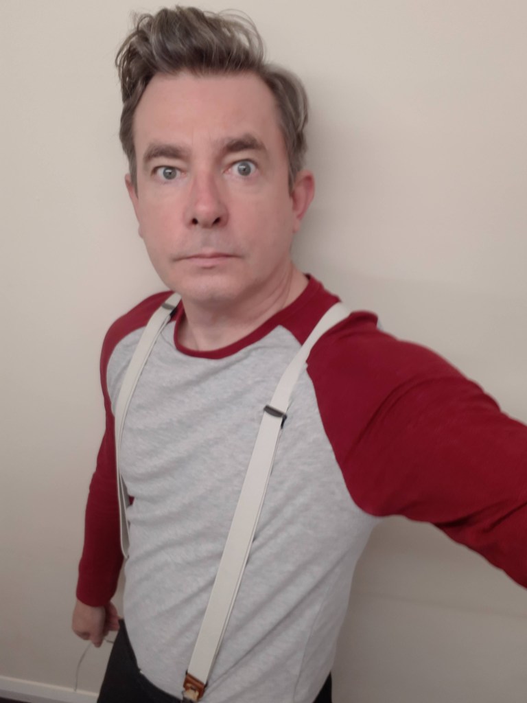 A man takes a selfie, he has wide eyes and is wearing suspenders.