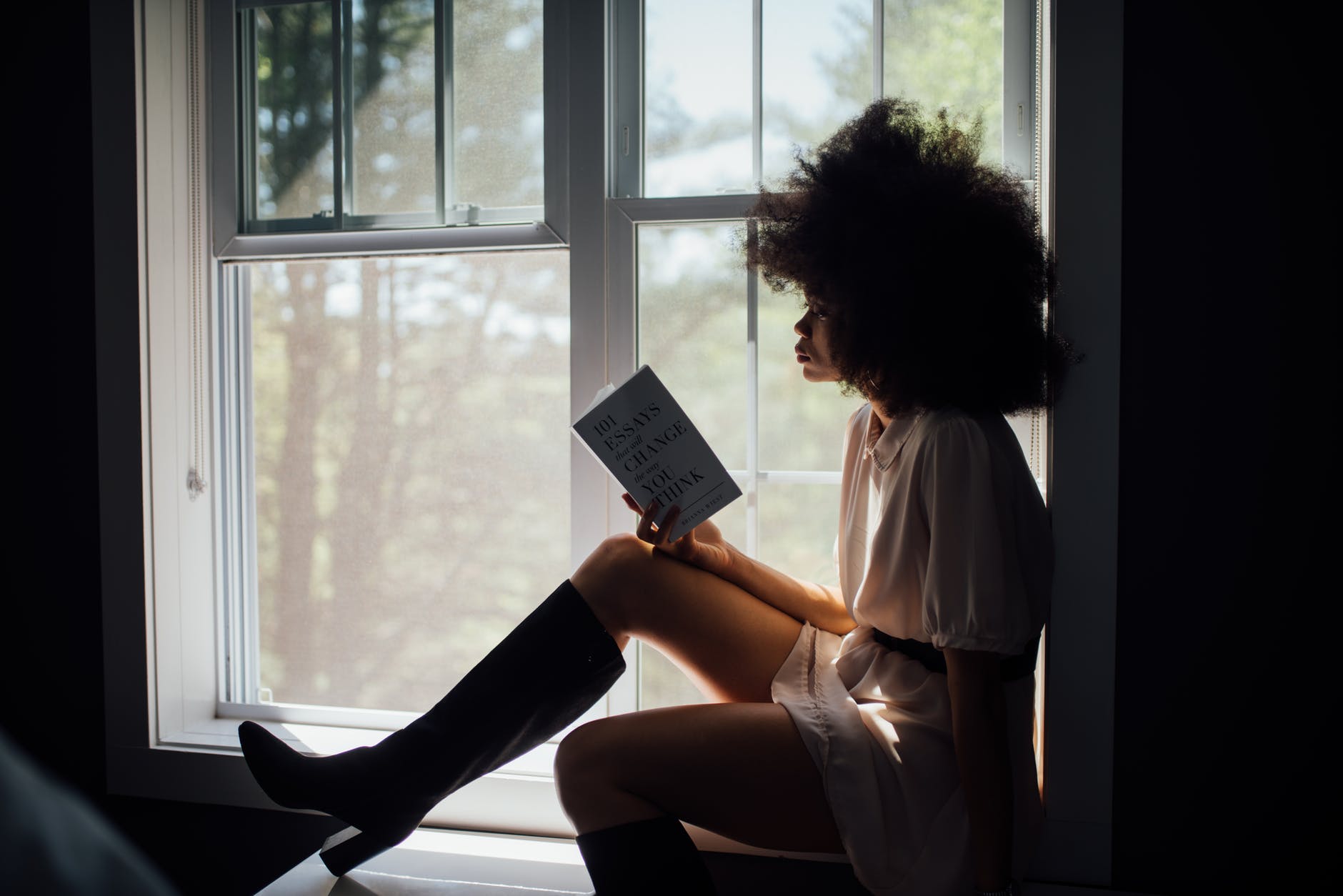 Woman sat on a windowsill reading a book. Her features are obscured by the light.