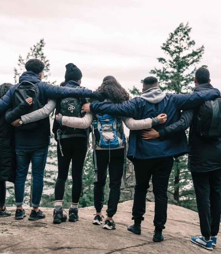 A group of friends stand on the edge of a forest, with their arms around each other. They have their backs to the camera
