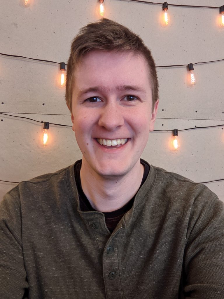 A portrait image of a young man with blond hair, smiling at the camera in front of fairy lights