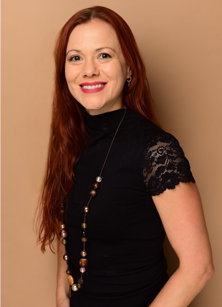 A woman smiles at the camera. She has red hair and wears a black lace top.