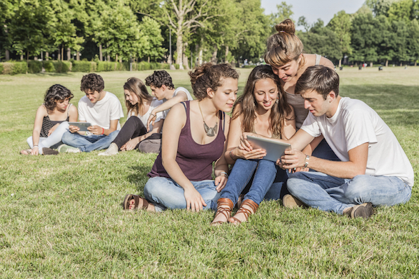 A group of friends sit on grass and look at a notepad or iPad