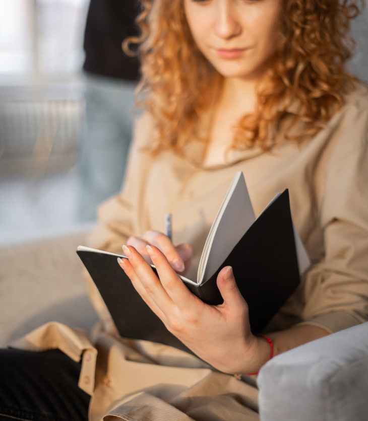 young woman writing in notebook in comfortable armchair- tricks to feel on track