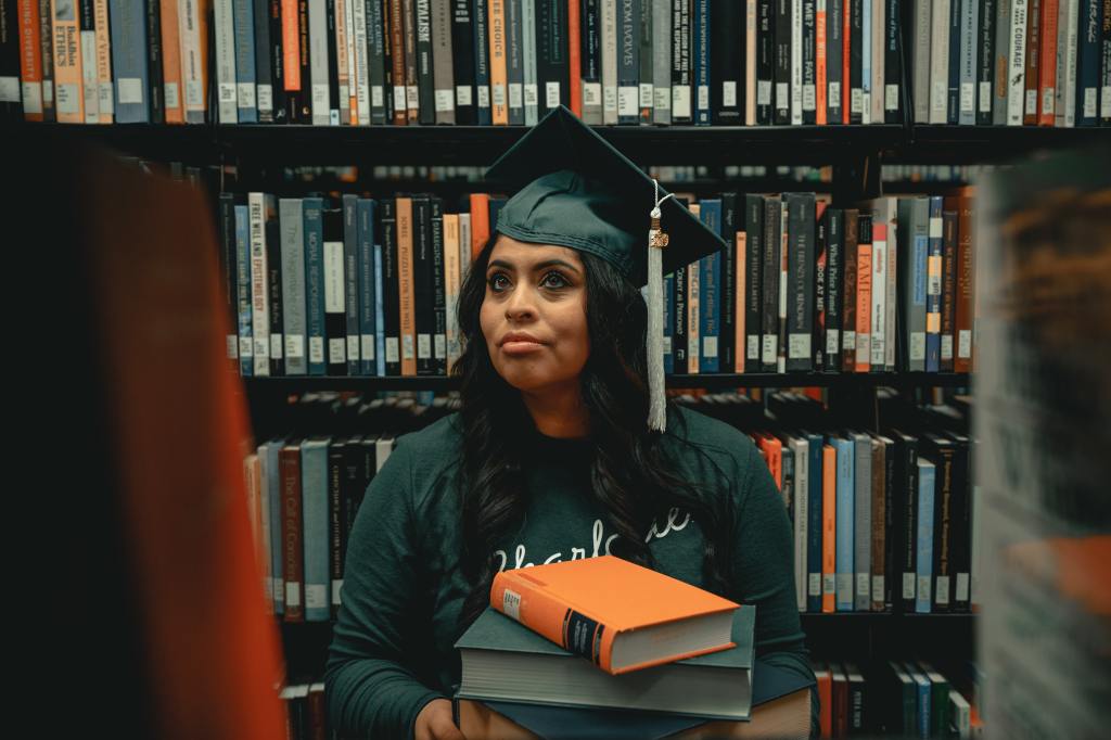 Woman wearing cap and gown in the library stacks, holding books- 8 things to do after you've finished University