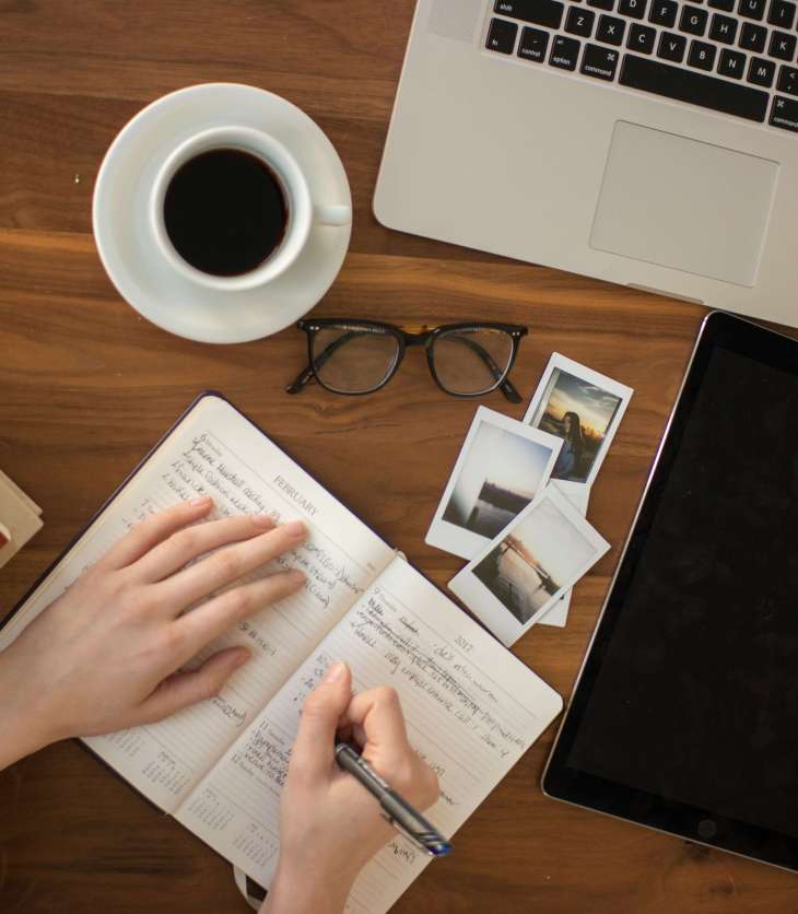 person writing in a notebook on a desk with laptop, coffee cip, polaroids, glasses, ipad and pile of books