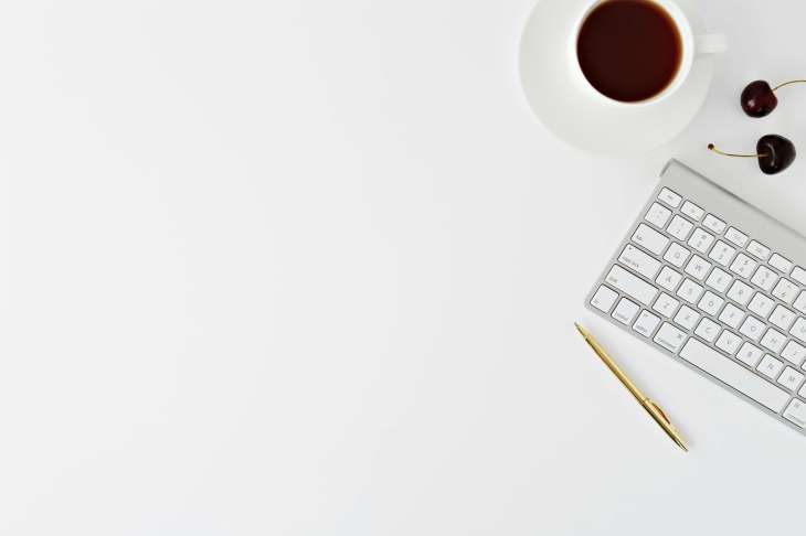 white desk with a cup of coffee, a keyboard, two cherries and pen at the right-hand side