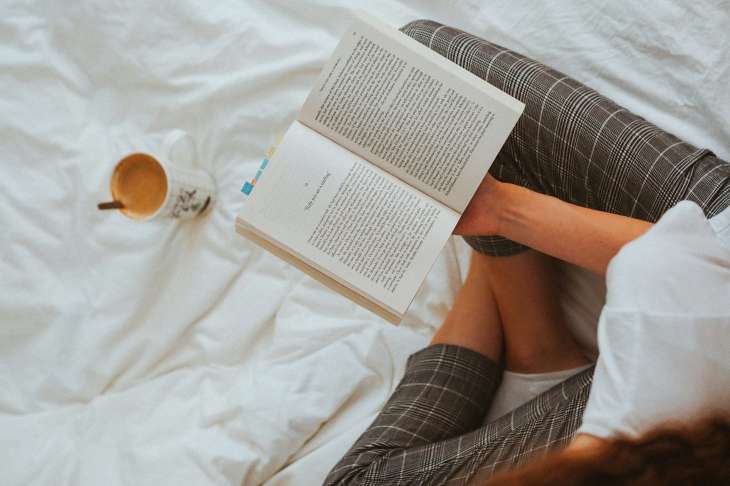 person holding a book open on a duvet with a teacup in front of them