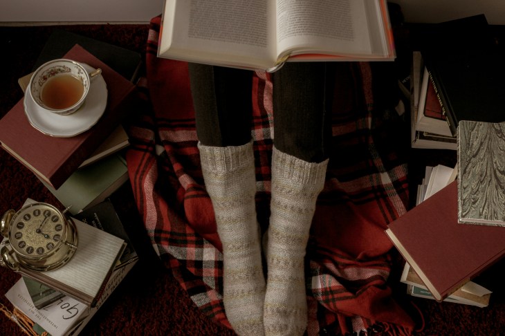 just a pair of legs wearing thick Autumn socks with a book on the lap and a Tartan blanket under the feet - pile of books around the legs and a teacup of tea