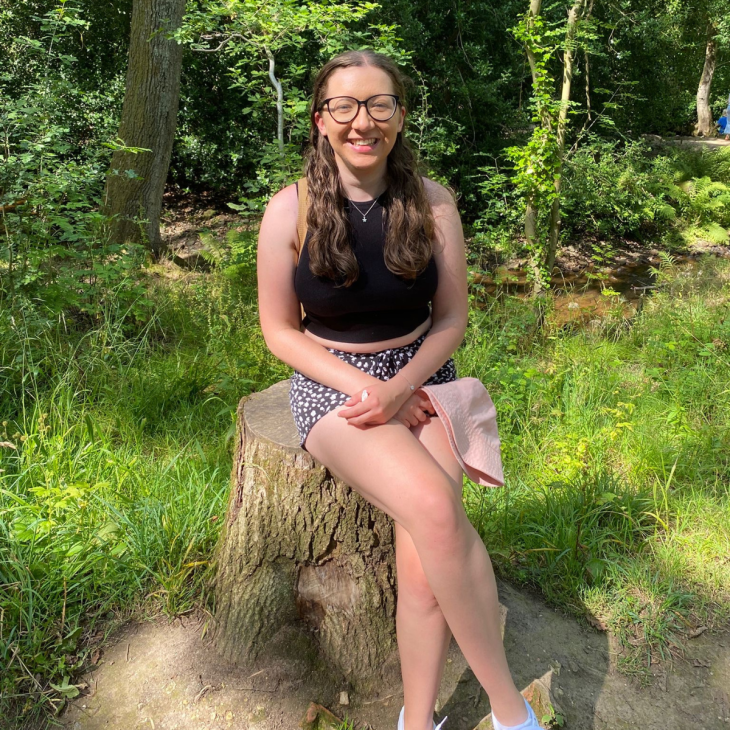 young woman sat on a tree stump with a pink hat in her hand smiling