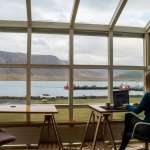 woman working with her back to the camera at a desk with a laptop - huge window in front of her with cliffs and body of water