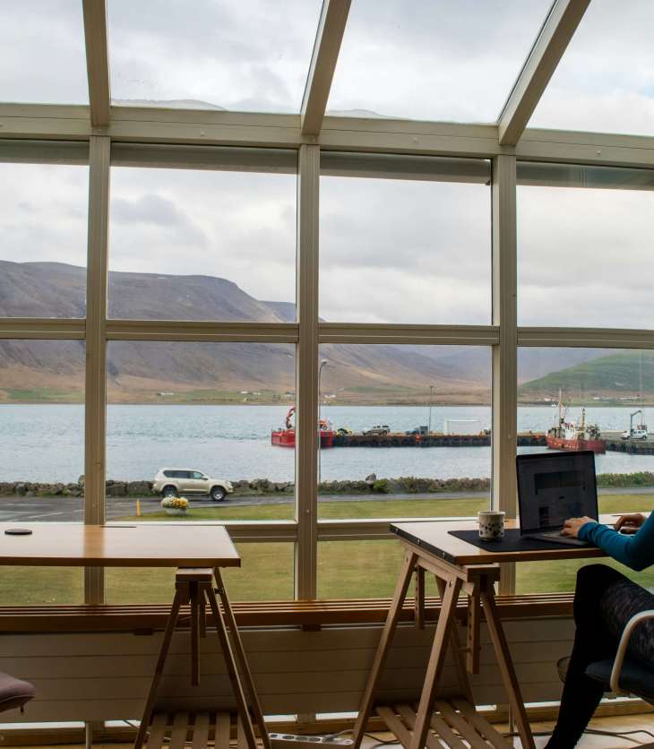 woman working with her back to the camera at a desk with a laptop - huge window in front of her with cliffs and body of water