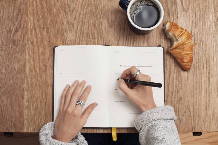 hands writing in a notebook on a table with a croissant (that has a bite out of it) and a mug of coffee