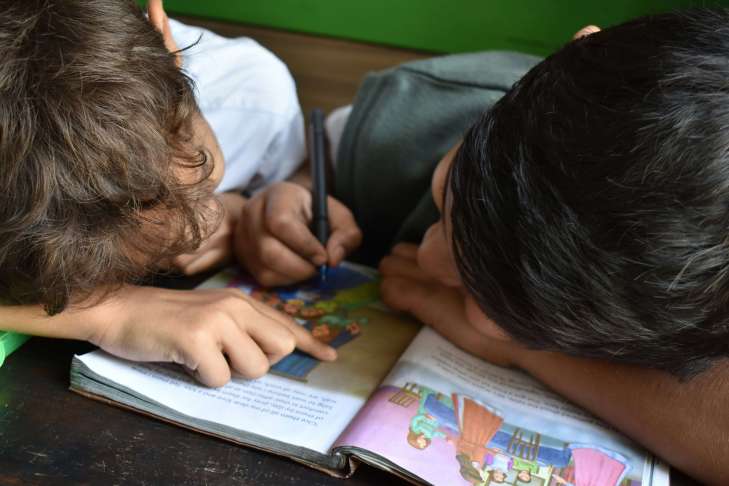 two kids leaning over a book on a desk and using a pen to write in it