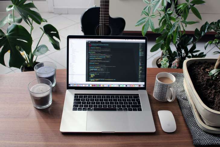 laptop on a desk surrounded by other items such as green plants, two candles, a mouse and a mug