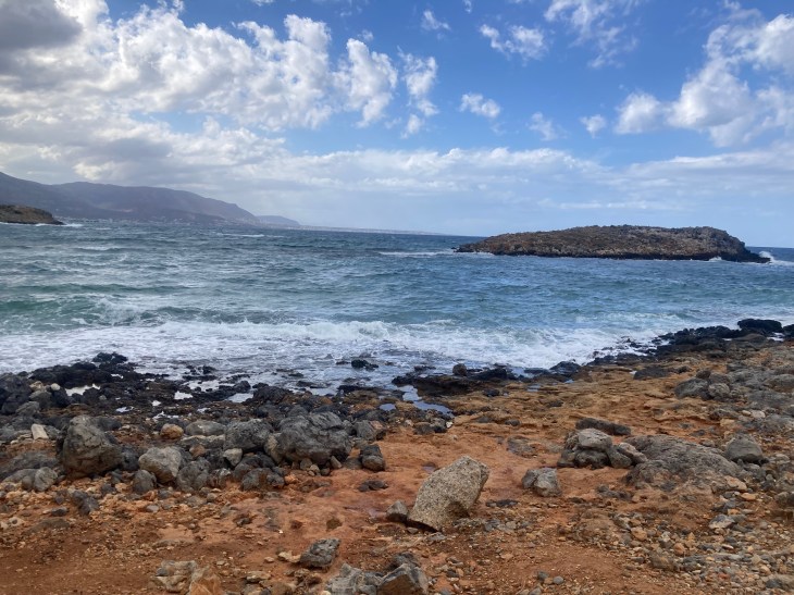 image of a rocky cliffside with the choppy sea in Crete - also a hill in the background