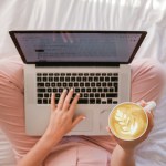 person sat cross-legged with a laptop on her knee and a cup of coffee in her right hand
