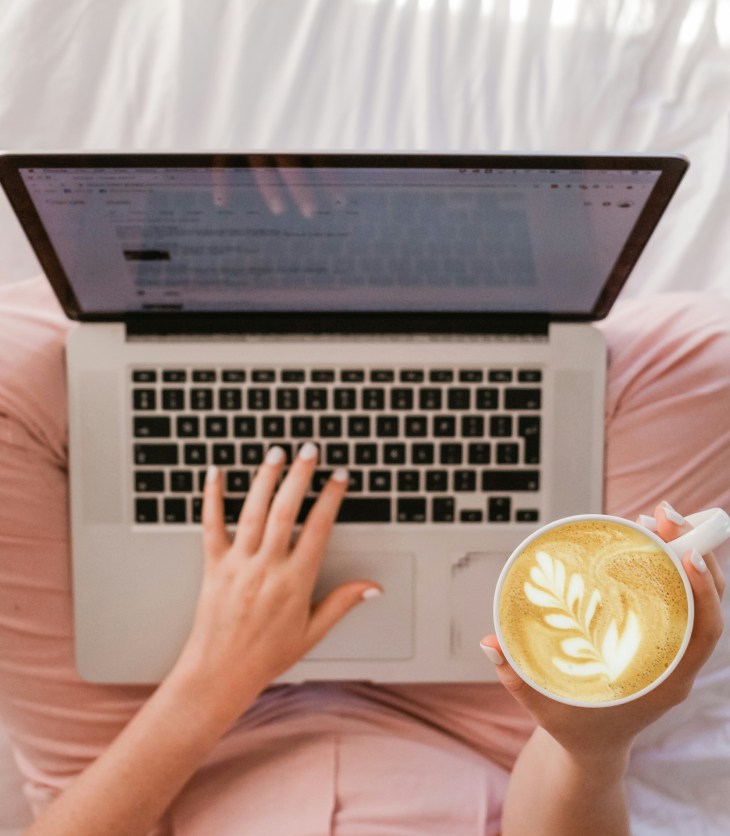 person sat cross-legged with a laptop on her knee and a cup of coffee in her right hand